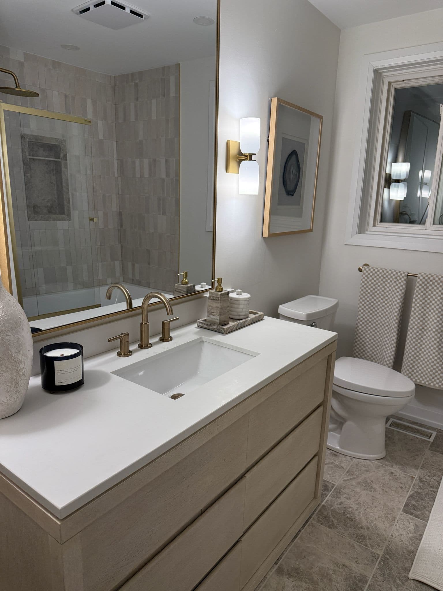 Bathroom renovation with light oak vanity, brass fixtures, large mirror, glass shower, and warm neutral tile floor