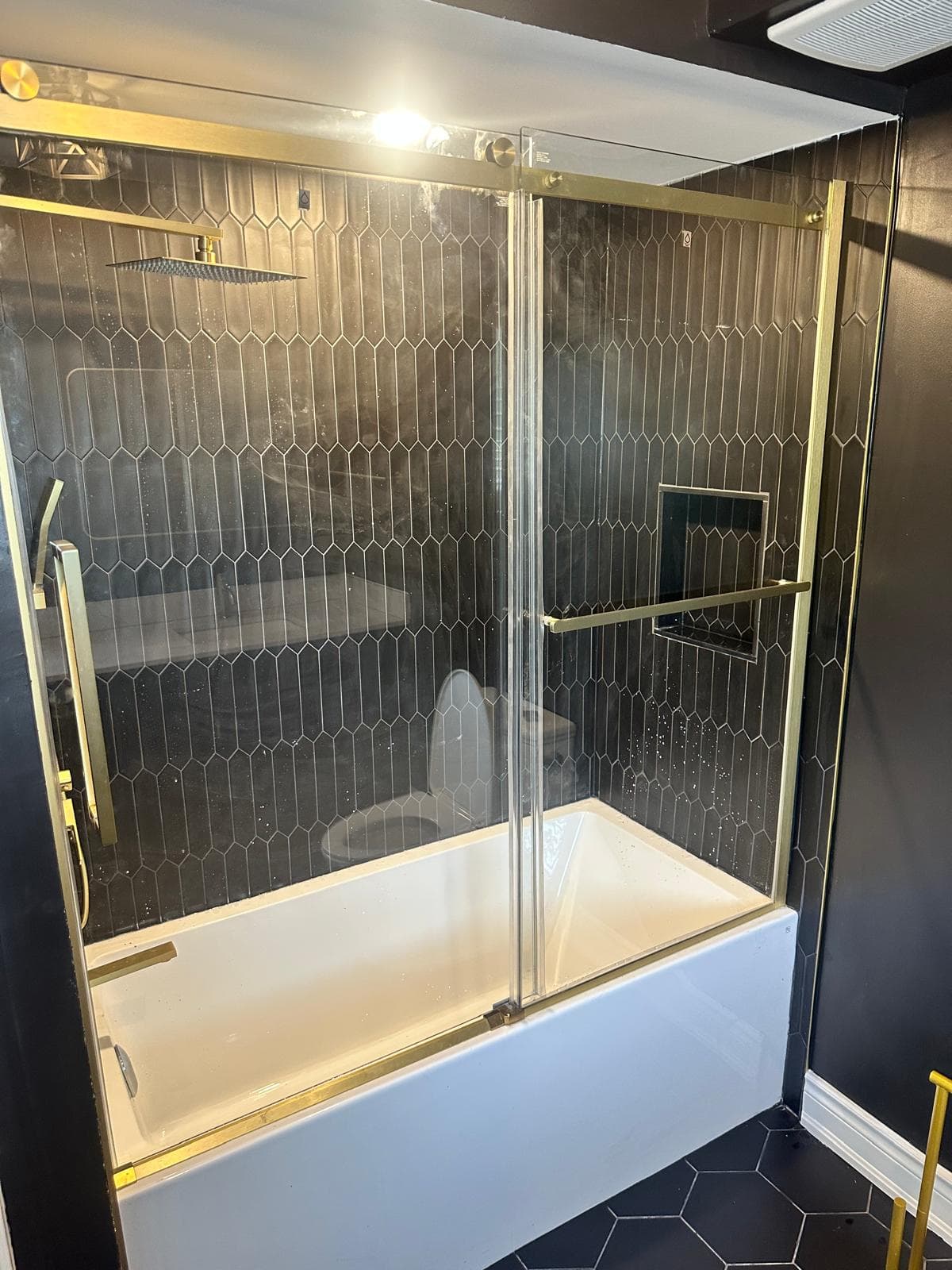 Tub-shower with black picket tile, rain shower head, and gold-framed sliding glass door